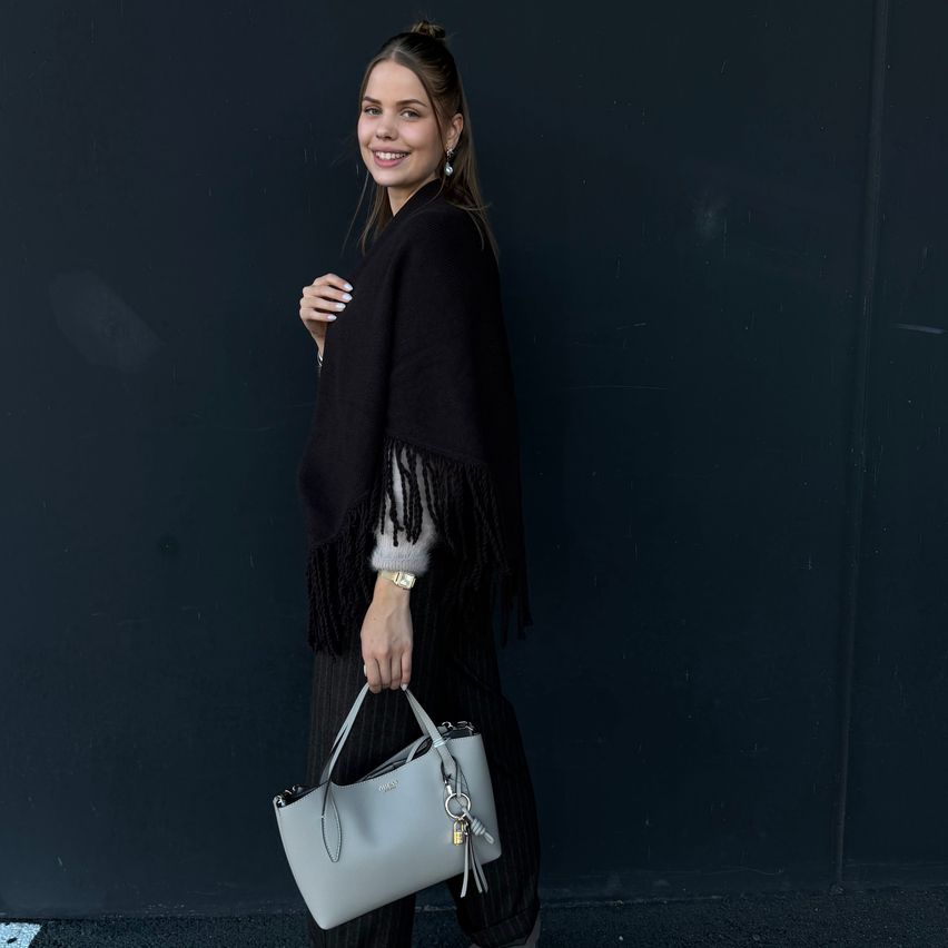 A woman in a black outfit with fringes and a gray handbag poses against a dark wall.