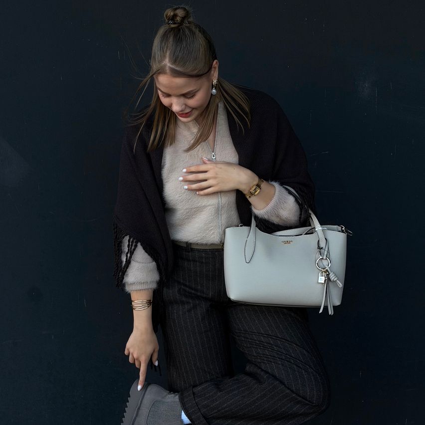 A woman wearing a white sweater, black jacket, and black pants is holding a grey handbag and smiling.