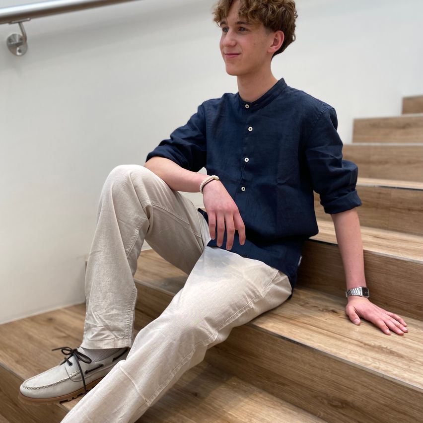 Ein junger Mann sitzt auf einer hölzernen Treppe, trägt ein marineblaues Hemd und beige Hose und posiert lächelnd für ein Foto.