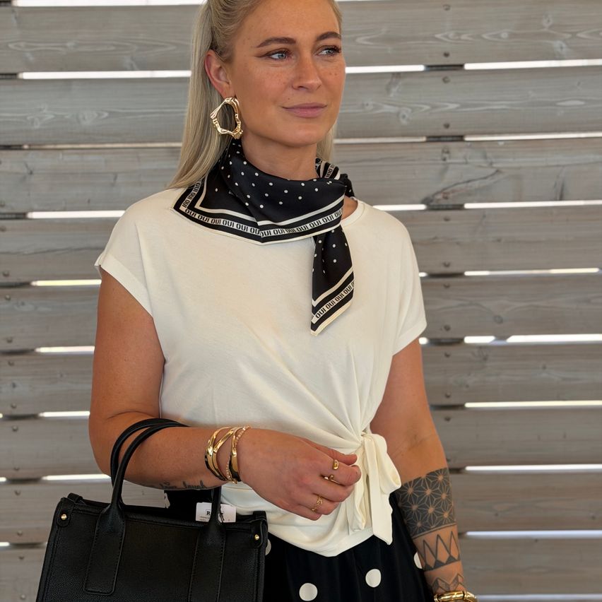 Eine Frau mit blonden Haaren trägt ein weißes Oberteil, einen schwarzen Punktschal und einen schwarzen Rock und hält eine schwarze Handtasche. Sie steht vor einer Holzwand.
