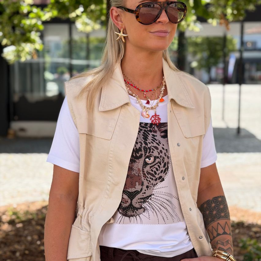 A woman with blonde hair stands in a park wearing a white shirt with a tiger print, a beige vest, and a necklace with a starfish pendant. She has tattoos on her arm.