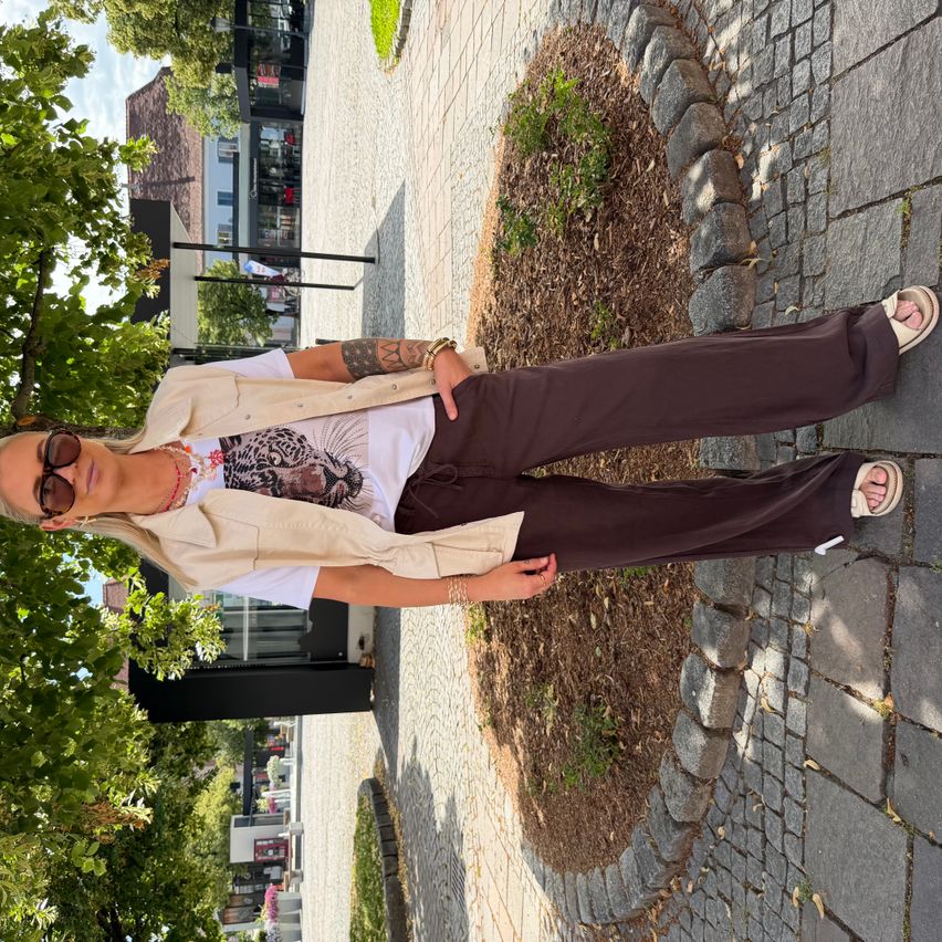 A woman in a white shirt with a tiger print and brown pants is standing on a brick pathway. She wears sunglasses and has a tattoo on her arm.