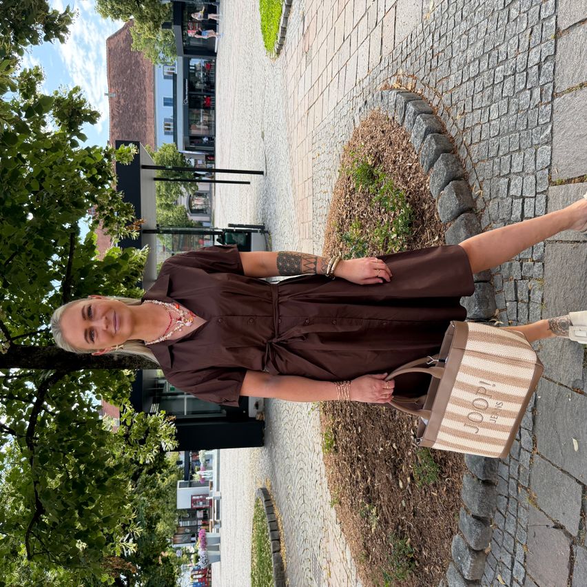 A woman with blonde hair is standing on a brick path, wearing a brown dress and holding a striped bag. Behind her, there are trees and buildings.