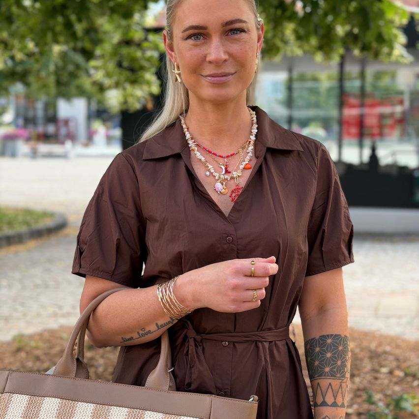 A woman wearing a brown dress and holding a handbag stands outdoors, smiling. She has tattoos on her arms and is wearing multiple necklaces. The background shows a street with buildings and trees.