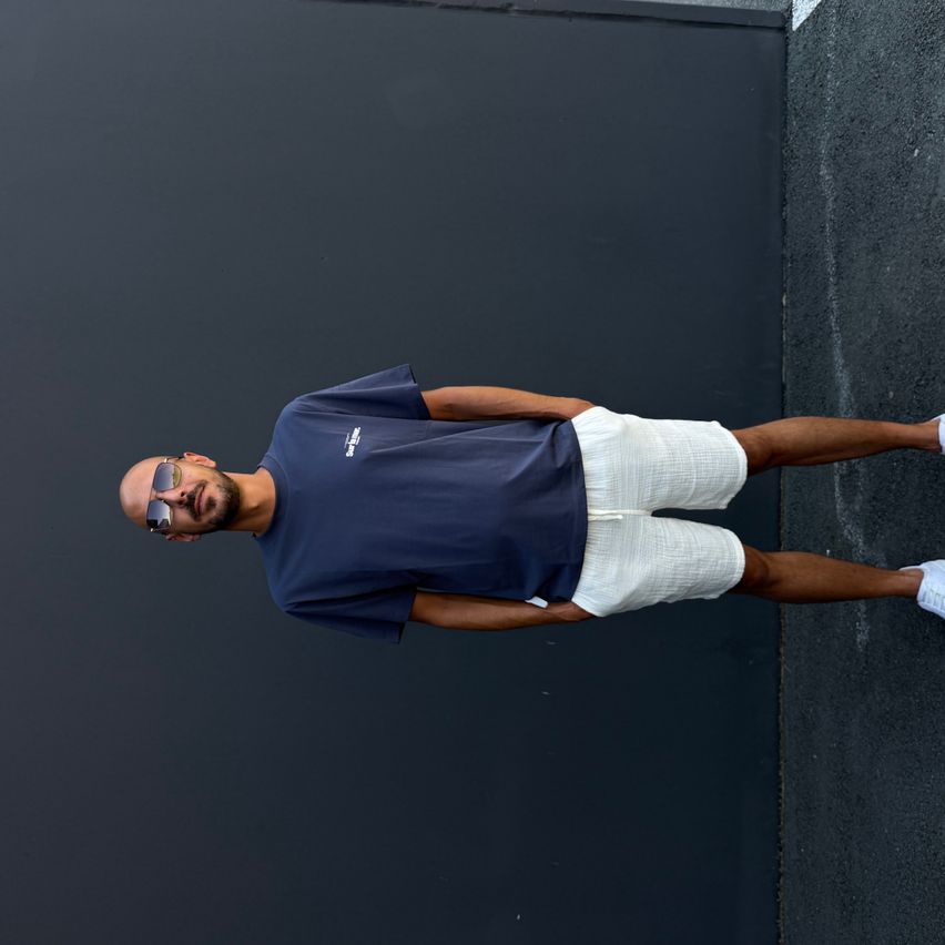 A man in a navy blue shirt and white shorts poses against a black wall. He wears sunglasses and white shoes.