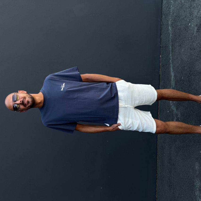 A man lies on a black wall, smiling. He wears a navy blue t-shirt and white shorts. He has a beard and sunglasses on.