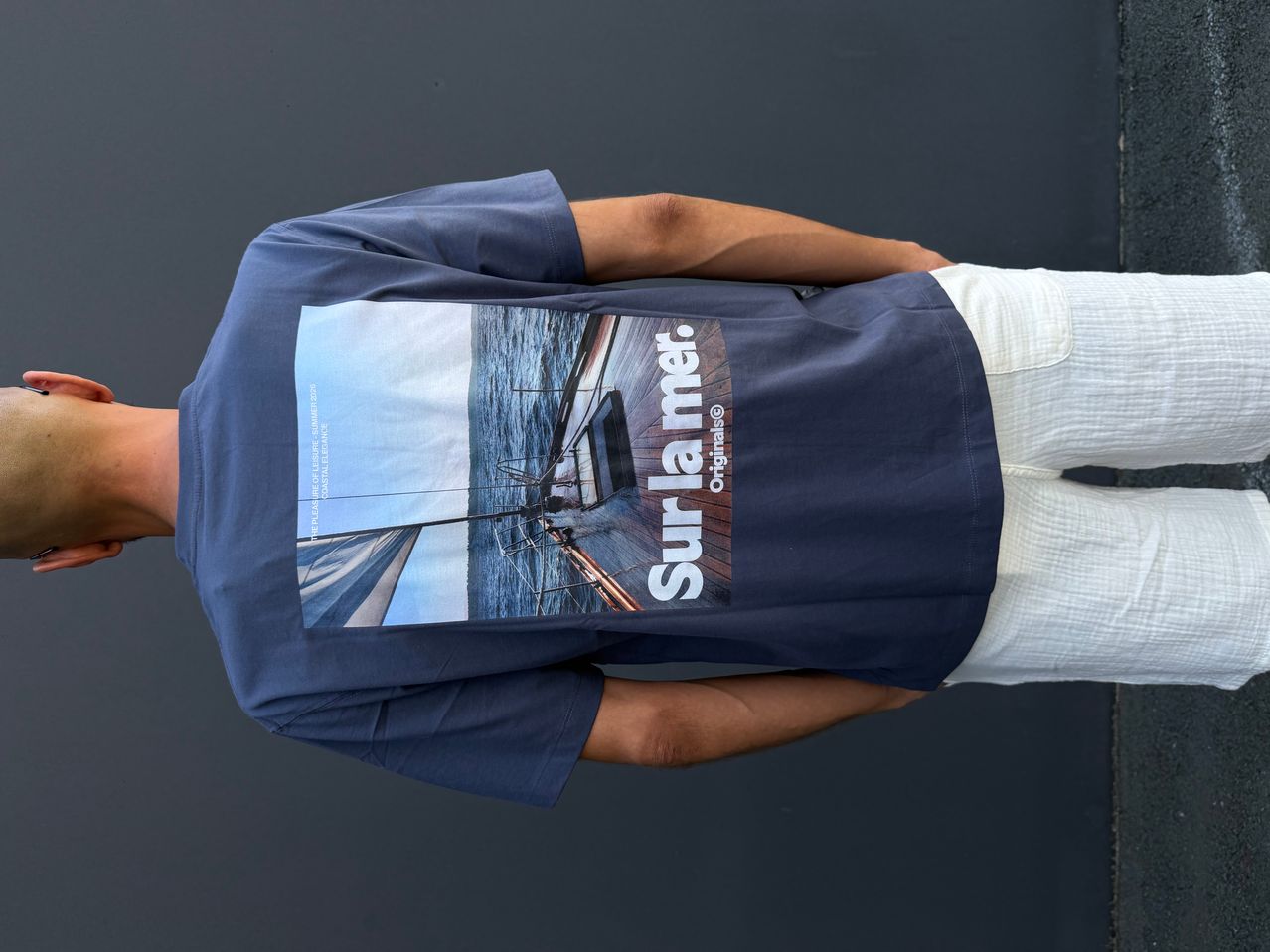 A person wears a navy blue t-shirt with a sailboat image. The shirt reads 'Surlamer' with a picture of a sailboat on the front.
