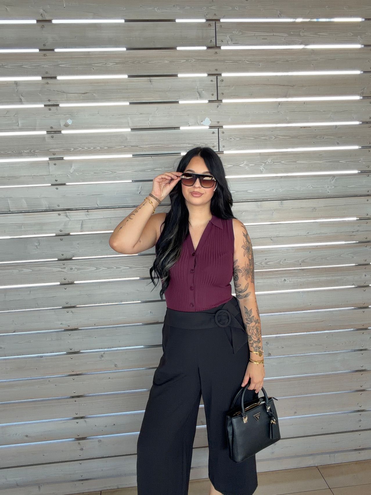 A woman with tattoos on her arms stands against a wooden wall, wearing a maroon top, black pants, and sunglasses, holding a black handbag.