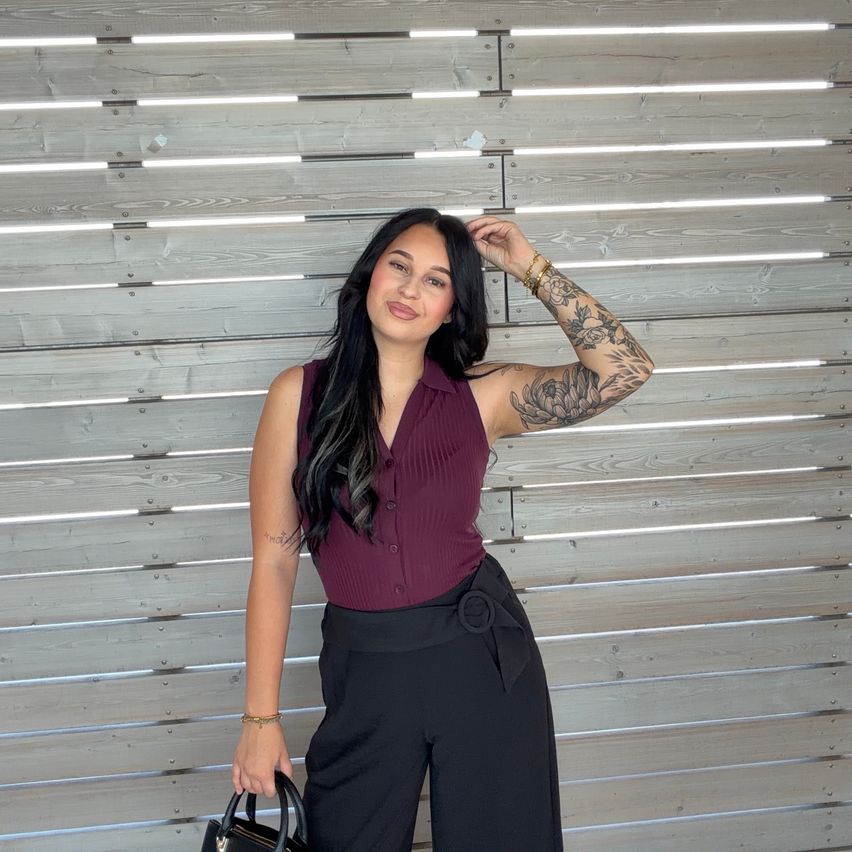 A woman with tattoos on her arms stands in front of a wooden wall, holding a handbag. She wears a maroon sleeveless top and black wide-legged pants.