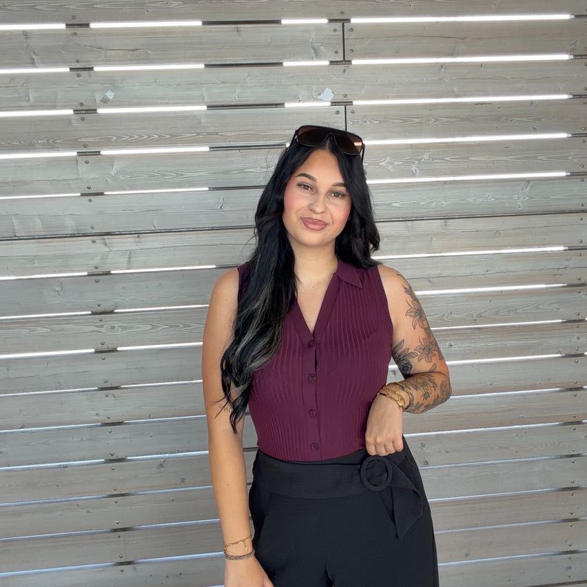 A woman with long black hair stands in front of a wooden wall, wearing a maroon sleeveless top and black pants. She has tattoos on her arms and is wearing sunglasses on her head.