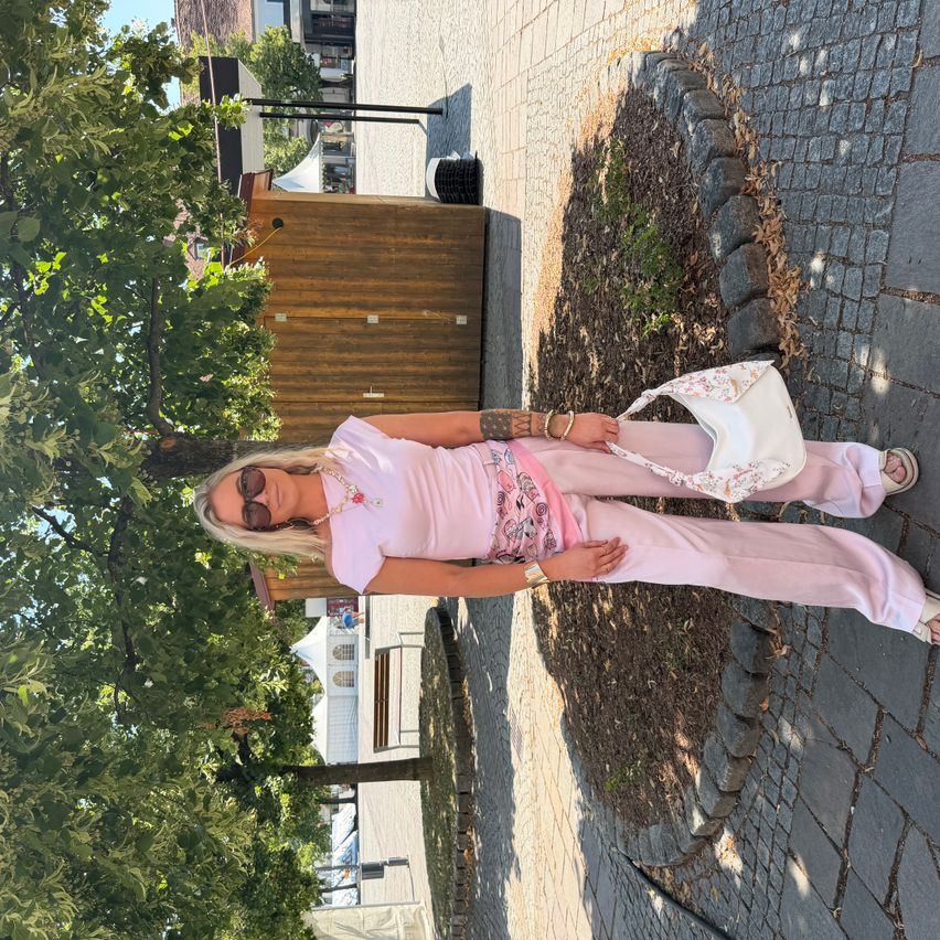 Eine Frau in rosa Kleidung posiert in einem Außenbereich mit einem Baum und gepflastertem Weg. Sie hält eine weiße Handtasche und steht neben einer Holzkonstruktion.