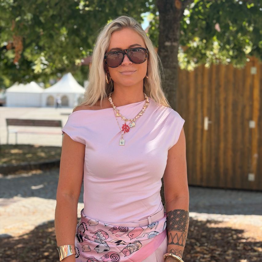 Eine Frau mit blonden Haaren und Sonnenbrille steht draußen. Sie trägt ein rosa Shirt und eine Halskette mit Blumenanhänger. Sie hat ein Tattoo am Arm und einen rosa Rock. Im Hintergrund befinden sich Bäume, ein Holzzaun und ein weißes Zelt.
