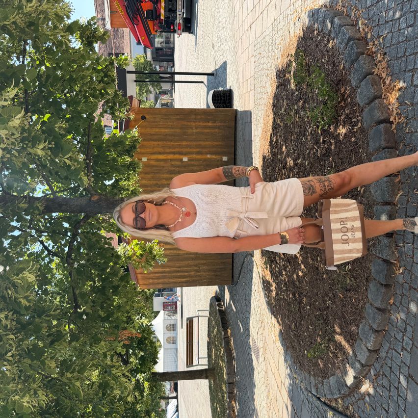 Eine Frau posiert mit einer Tasche in der Hand unter einem Baum an einem sonnigen Tag.