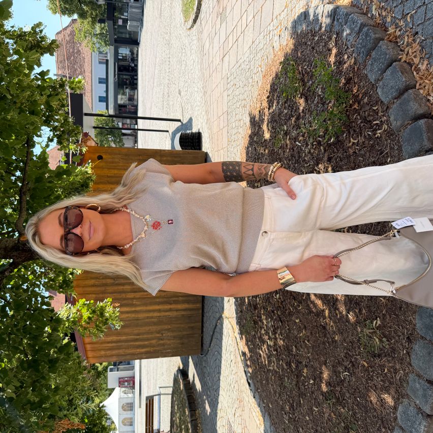 A woman in a light gray top and white pants poses outdoors under a tree, wearing sunglasses and holding a bag. Behind her is a wooden fence, and in the distance are buildings.