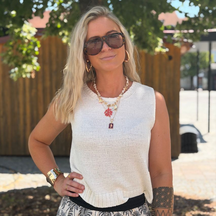 A woman with blonde hair, wearing sunglasses, a white sleeveless top, and a necklace. She has a tattoo on her right arm and a black belt around her waist.