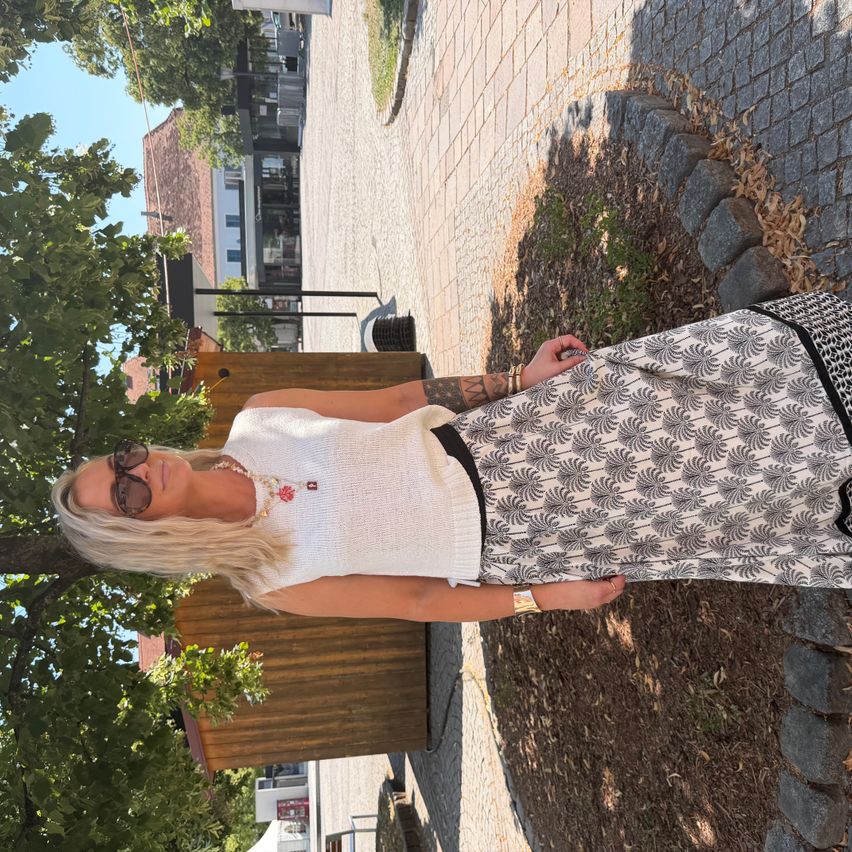 A blonde woman in a white top and black skirt stands beneath a tree, near a wooden fence. Buildings and greenery are in the background.
