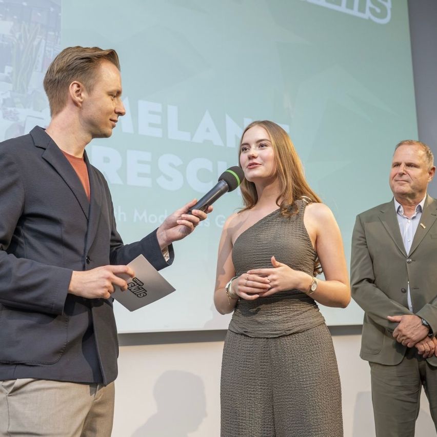 A woman in a sleeveless dress stands between two men, one holding a microphone and the other with folded hands, in front of a projection screen.