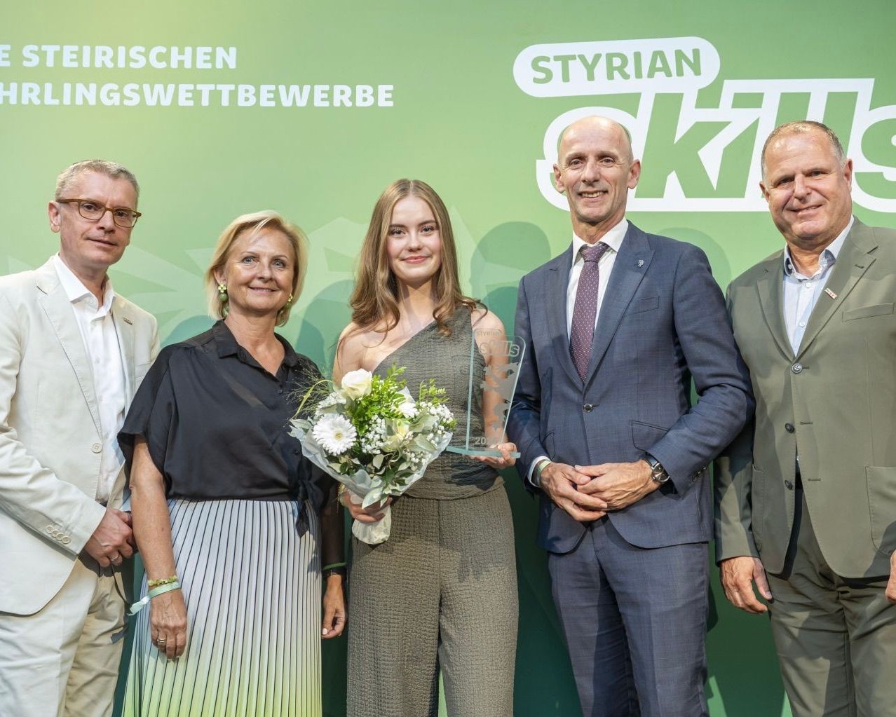 Five individuals stand in front of a green backdrop with 'Styrian Ski' text. A woman in the center holds a bouquet and an award. The man on the right wears a watch and tie.
