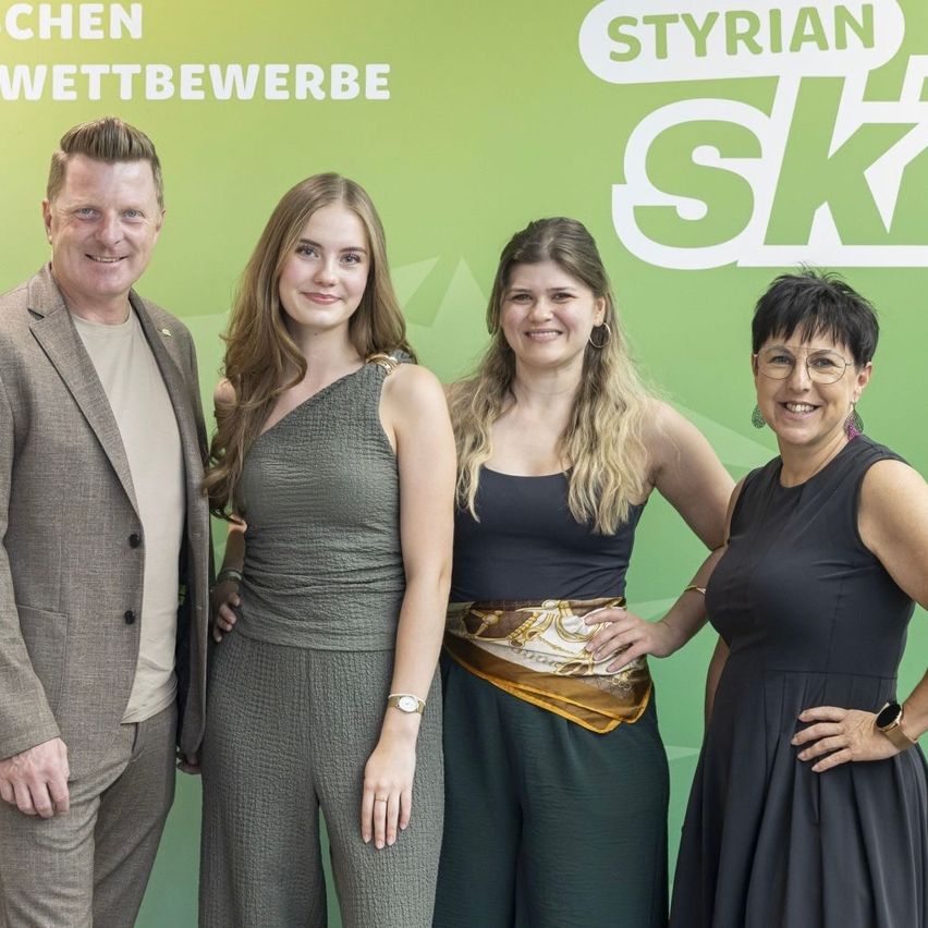 Four individuals stand together in front of a green backdrop with the word 'Styrian' and 'Ski' written in white. The man on the left wears a suit, while the women wear casual dresses. They all appear to be smiling.