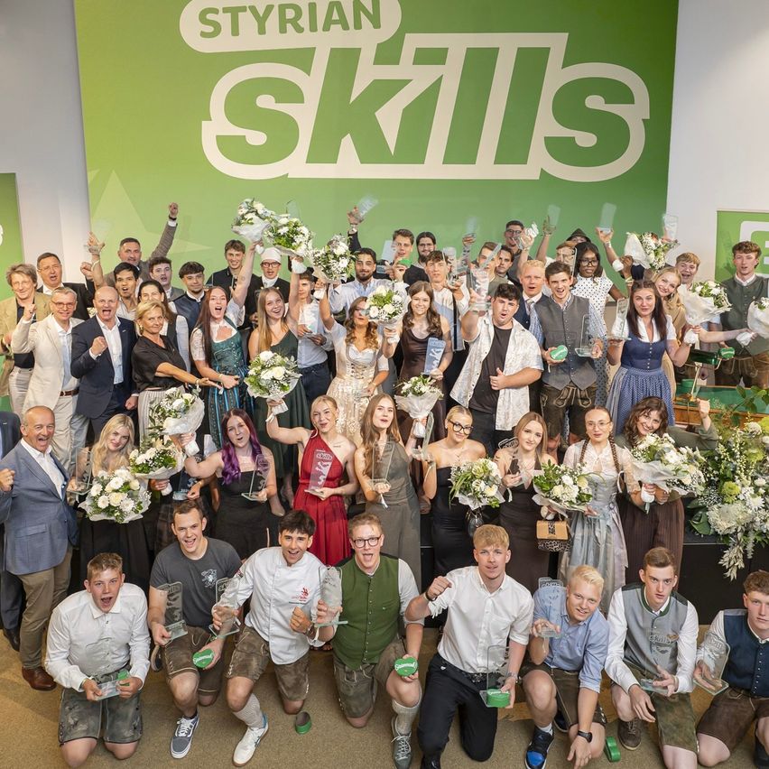 A large group of people dressed in traditional attire and holding flowers and awards pose for a photo in front of a green banner that reads 'Styrian Skills'.