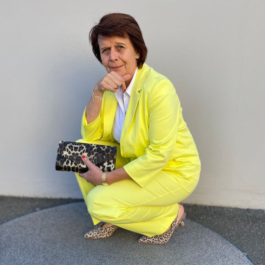 A woman in a bright yellow suit and leopard print shoes holds a matching clutch bag, posing against a white wall.