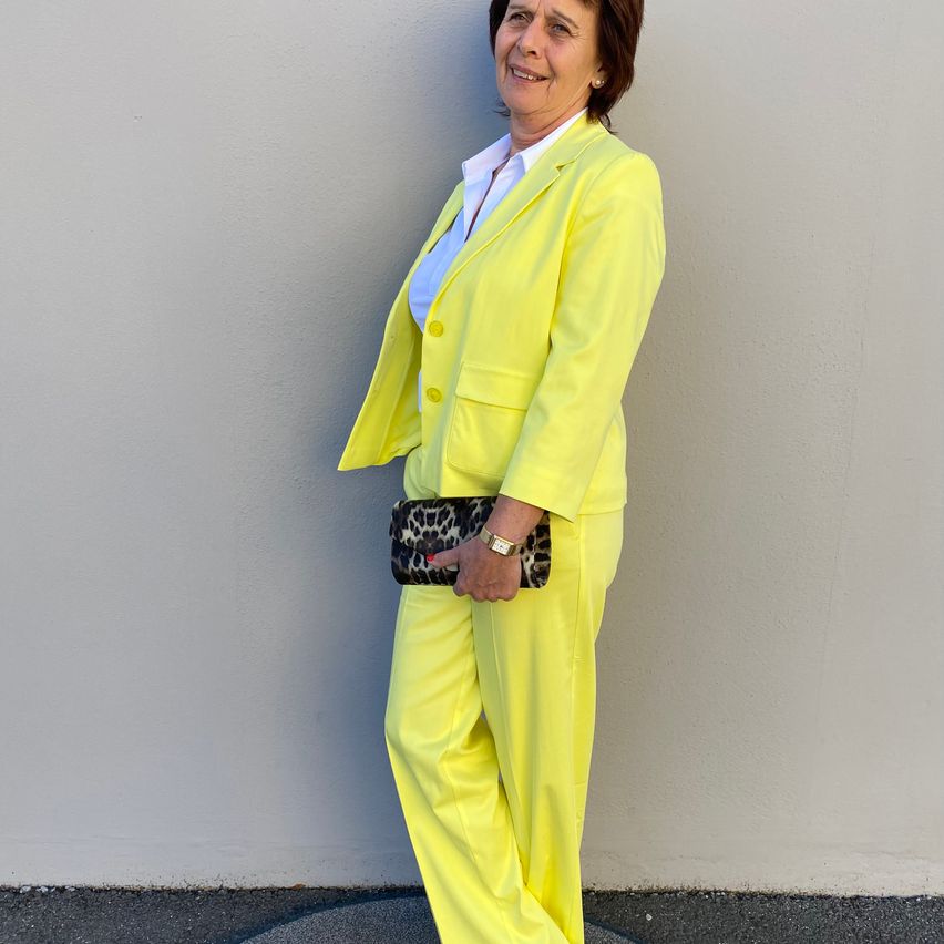 A woman in a bright yellow suit stands against a white wall, smiling and holding a leopard print clutch.