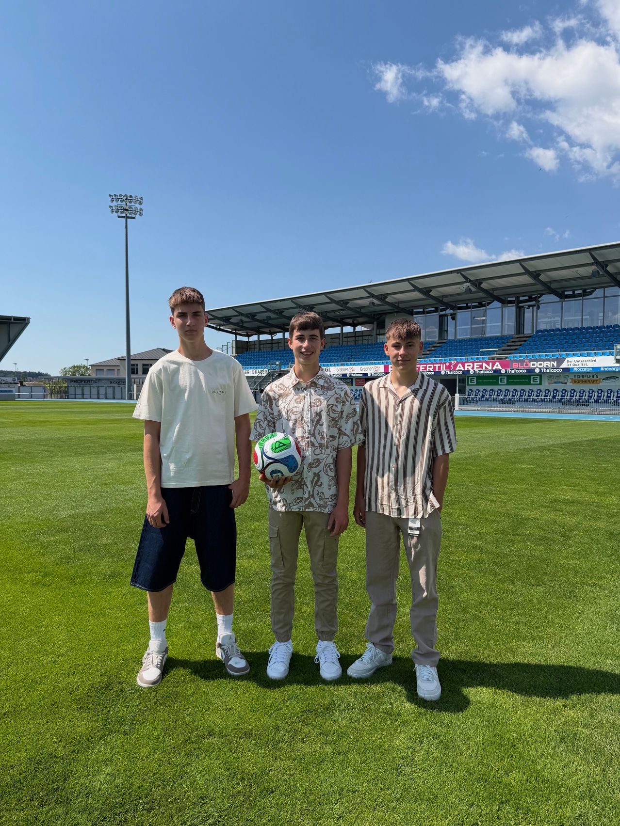 Three young men stand on a soccer field, one holding a soccer ball. They are dressed casually and appear to be posing for a photo. The field is surrounded by empty bleachers.