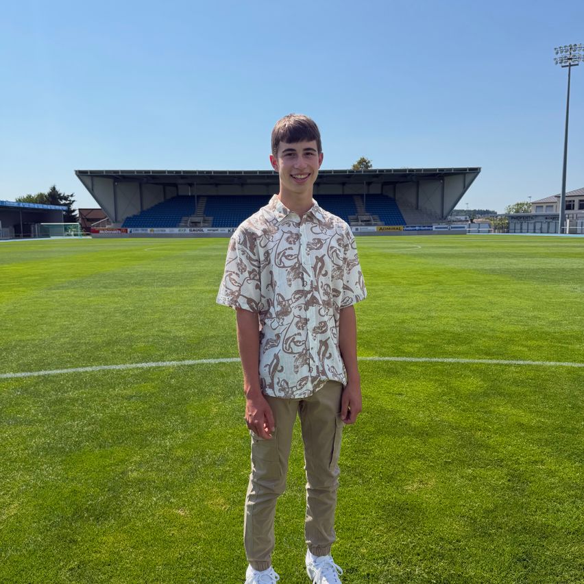 A young man stands on a soccer field, smiling, with an empty stadium in the background.