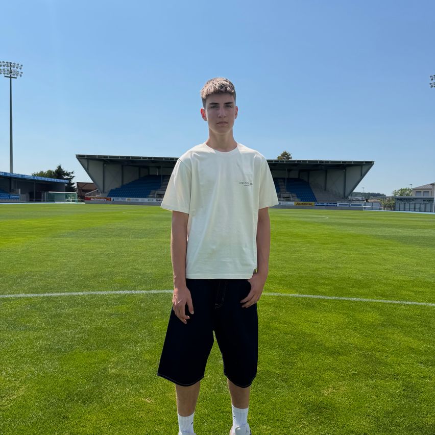 A young man stands on a soccer field, wearing a white t-shirt and black shorts. He is looking to the left, with a stadium and bleachers in the background.