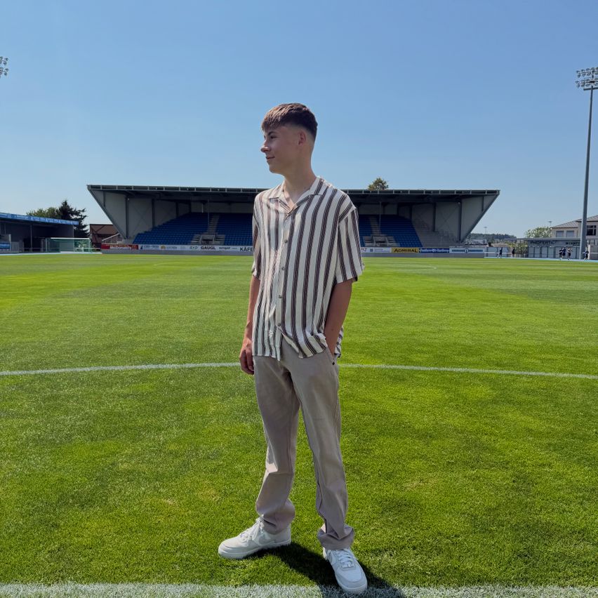 A young man stands on a soccer field, wearing a striped shirt and khaki pants. He is looking to the right, with an empty stadium in the background.