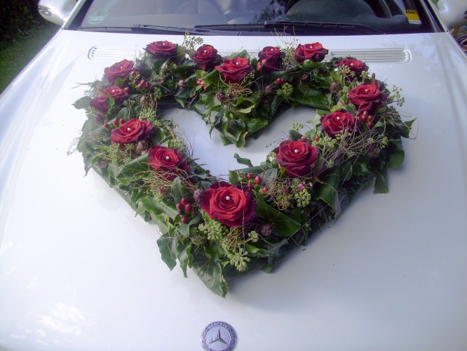 Ein Mercedes-Auto, geschmückt mit einer herzförmigen Anordnung aus roten Rosen und grünen Blättern.
