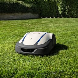 Ein weißer und blauer robotergesteuerter Rasenmäher steht auf einem grünen Rasen mit einer Steinmauer und Hecken im Hintergrund.