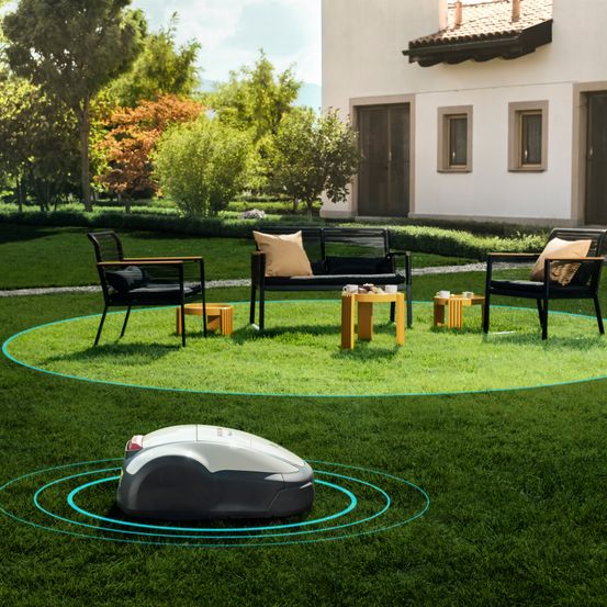 Ein Rasenmähroboter steht auf einem kreisförmigen Rasen mit einem Haus im Hintergrund. Drei schwarze Stühle mit Kissen umgeben einen runden Holztisch.