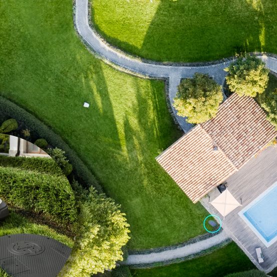 Luftaufnahme eines Hauses mit Ziegeldach, Swimmingpool und Gehweg. Der Rasen ist gut gepflegt, und es gibt Bäume und Sträucher an der Seite. Ein kreisförmiges Objekt ist hervorgehoben.