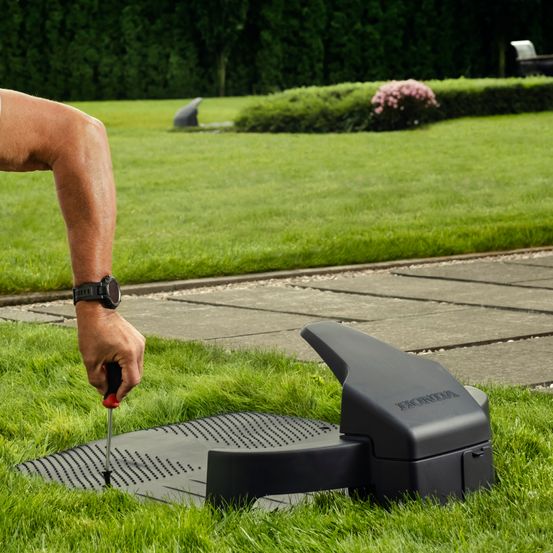 Eine Person hält einen Schraubendreher in der Nähe eines schwarzen Honda-Rasenmähers in einem grünen Garten.