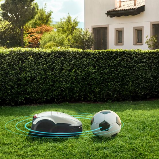 Ein robotergesteuerter Rasenmäher ist auf dem Rasen, mit einem Fußball in der Nähe. Hinter dem Rasen steht eine Hecke und ein Haus mit einem Ziegeldach.