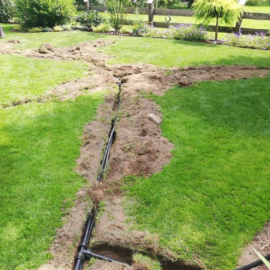 Ein Garten mit frisch verlegten schwarzen Rohren in der Mitte des Rasens. Der Boden ist teilweise entfernt, und es gibt Pflanzen und Büsche im Hintergrund.