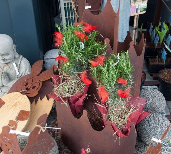Dekorative Ausstellung mit Topfpflanzen und Skulpturen in einem Gartenbereich. Eine Buddha-Statue und eine Hasenfigur stehen neben Eisenpflanzern mit roten Federn und Grün.