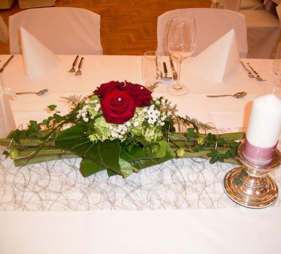 Ein Tisch mit einem Blumenarrangement aus roten Rosen und Grünpflanzen, einer Kerze und einer Tischdecke. Leere Stühle umgeben den Tisch.