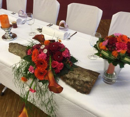 Ein langer Tisch mit weißen Stühlen und Blumen, einer Kerze und Weingläsern. Auf dem Tisch liegt ein Baumstamm.