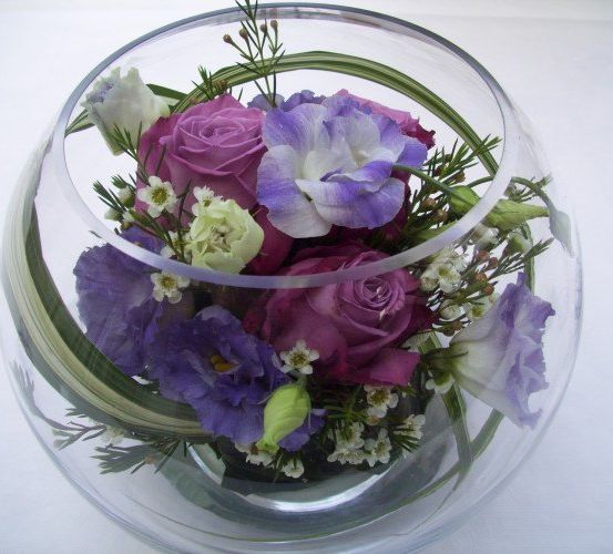 Ein durchsichtiger Glasteller enthält ein farbenfrohes Blumenarrangement. Die Blumensträuße bestehen aus violetten und rosa Rosen, weißen und violetten Schwertlilien und verschiedenen grünen Blättern. Die Blumen sind in einem kreisförmigen Muster angeordnet, wobei einige Blütenblätter die Glasoberfläche berühren.