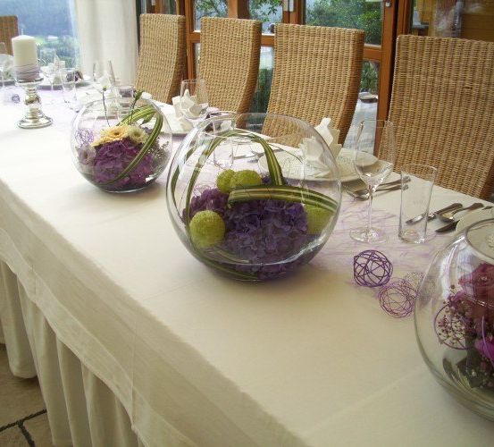 Ein langer Esstisch ist für eine Mahlzeit vorbereitet, mit drei Glasschüsseln gefüllt mit Blumen, umgeben von Stühlen und einem Blick auf einen Garten.