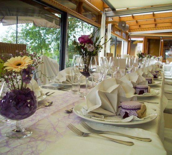 Ein wunderschön gedeckter Esstisch mit lila Blumen, gefalteten Servietten und Weingläsern. Der Tisch ist mit einer weißen Tischdecke bedeckt und von Stühlen umgeben.