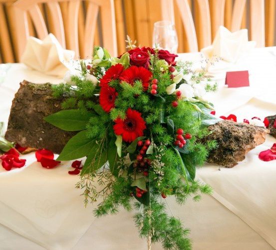 Ein Tisch für eine formelle Veranstaltung ist mit einer Tischdekoration aus roten Rosen, grünen Blättern und kleinen Steinen geschmückt. Er ist mit verstreuten Rosenblättern verziert und mit Weingläsern und gefalteten Servietten ausgestattet.