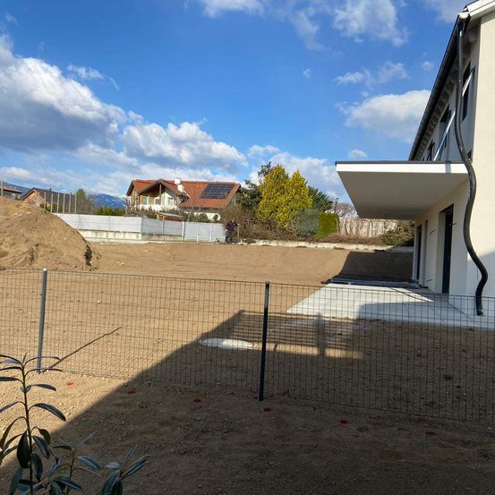 Ein eingezäunter Bauplatz mit einem im Hintergrund befindlichen Haus im Bau, sonniges Wetter und einem Haufen Erde.