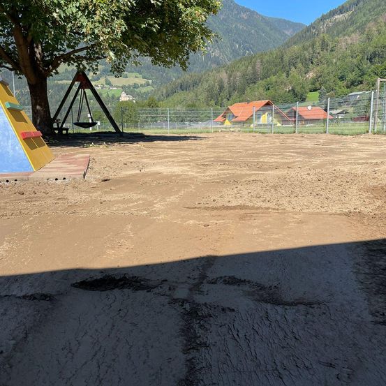 Ein leerer Spielplatz mit einem Schaukelgestell in der Nähe eines Baumes, umgeben von einem Zaun, mit Blick auf ein Haus und Berge in der Ferne.