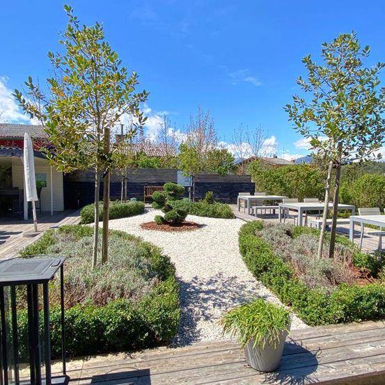 Ein Außenbereich mit Tischen und Stühlen, Kieswegen und gut geschnittenen Büschen. Hohe Bäume und eine Bank sind im Hintergrund zu sehen.