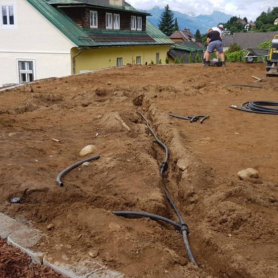 Ein Baustelle mit Arbeitern, die Kabel verlegen. Die Baustelle hat ein Haus mit grünem Dach. Bäume und ein Schubkarren sind im Hintergrund zu sehen.