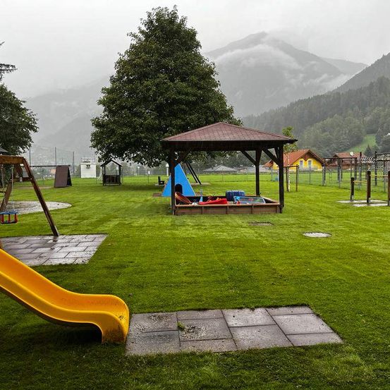 Ein Spielplatz mit Rutsche, Pavillon und anderem Equipment auf einer Rasenfläche mit Bergen im Hintergrund.