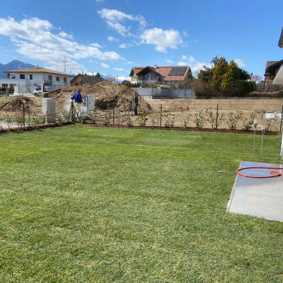 Ein Außenbereich mit grünen Rasenflächen und einem Zaun, zwei Personen arbeiten auf dem Land, eine steht auf einem Haufen Erde und die andere auf einem Haufen Schutt. In der Ferne stehen Häuser mit Solarmodulen auf den Dächern.